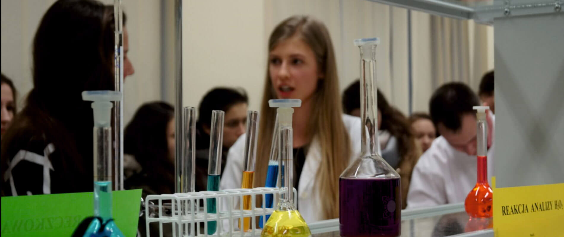 Zdjęcie przedstawia lekcję chemii. Na pierwszym planie próbówki z kolorowymi płynami, a w tle ucznionwie.
